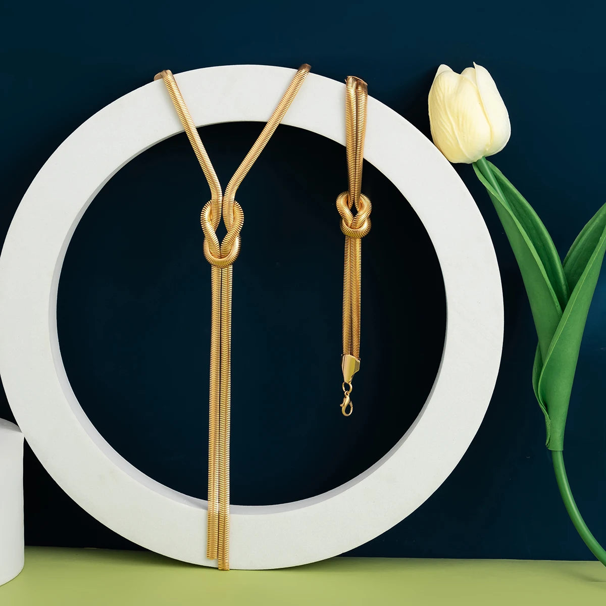 Gold necklaces on a white circular stand with a white tulip on a dark background. Women’s vintage goth snake twisted chain necklace & bracelet set with tassel, Y2K style jewelry accessories for fashion-forward looks.