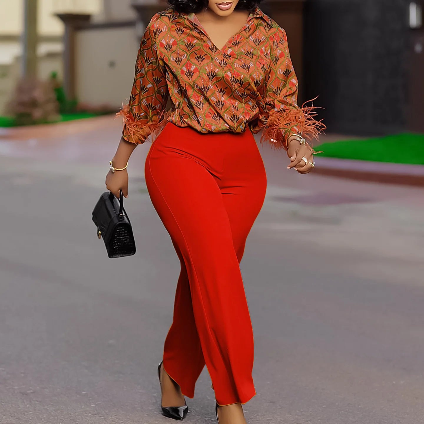 Woman in a patterned orange blouse and red pants walking outdoors. African print two-piece polyester outfit with satin printed shirt and high waist wide leg pants. Comfortable, elegant office wear for women.