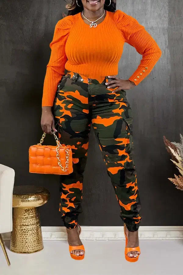 Person wearing an orange top and camouflage pants, holding an orange handbag against a dark wall.