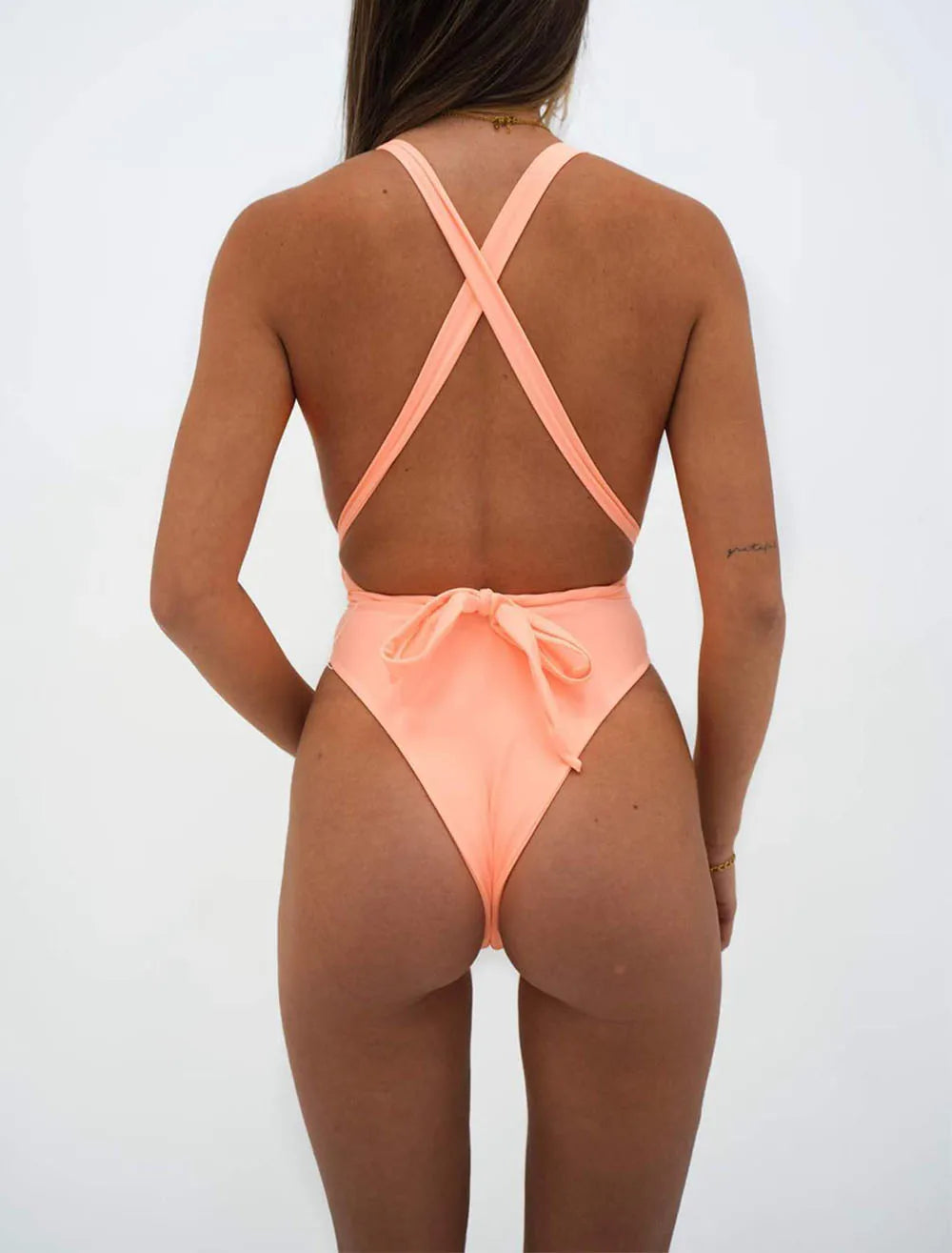 Person wearing a peach-colored swimsuit with criss-cross straps on a white background. Solid pink high cut v-neck monokini with lace-up detail, adjustable straps, beachwear for women, true to size, flattering, UK swim style.
