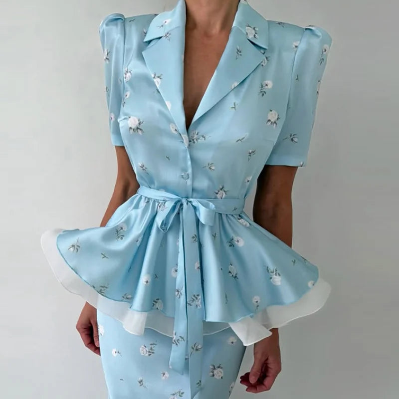 A light blue short-sleeve satin-like top with a small white floral print, notched lapels, a tie waist, and a flared peplum hem with white undershadow, shown on a model against a plain light background.