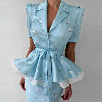 A light blue short-sleeve satin-like top with a small white floral print, notched lapels, a tie waist, and a flared peplum hem with white undershadow, shown on a model against a plain light background.