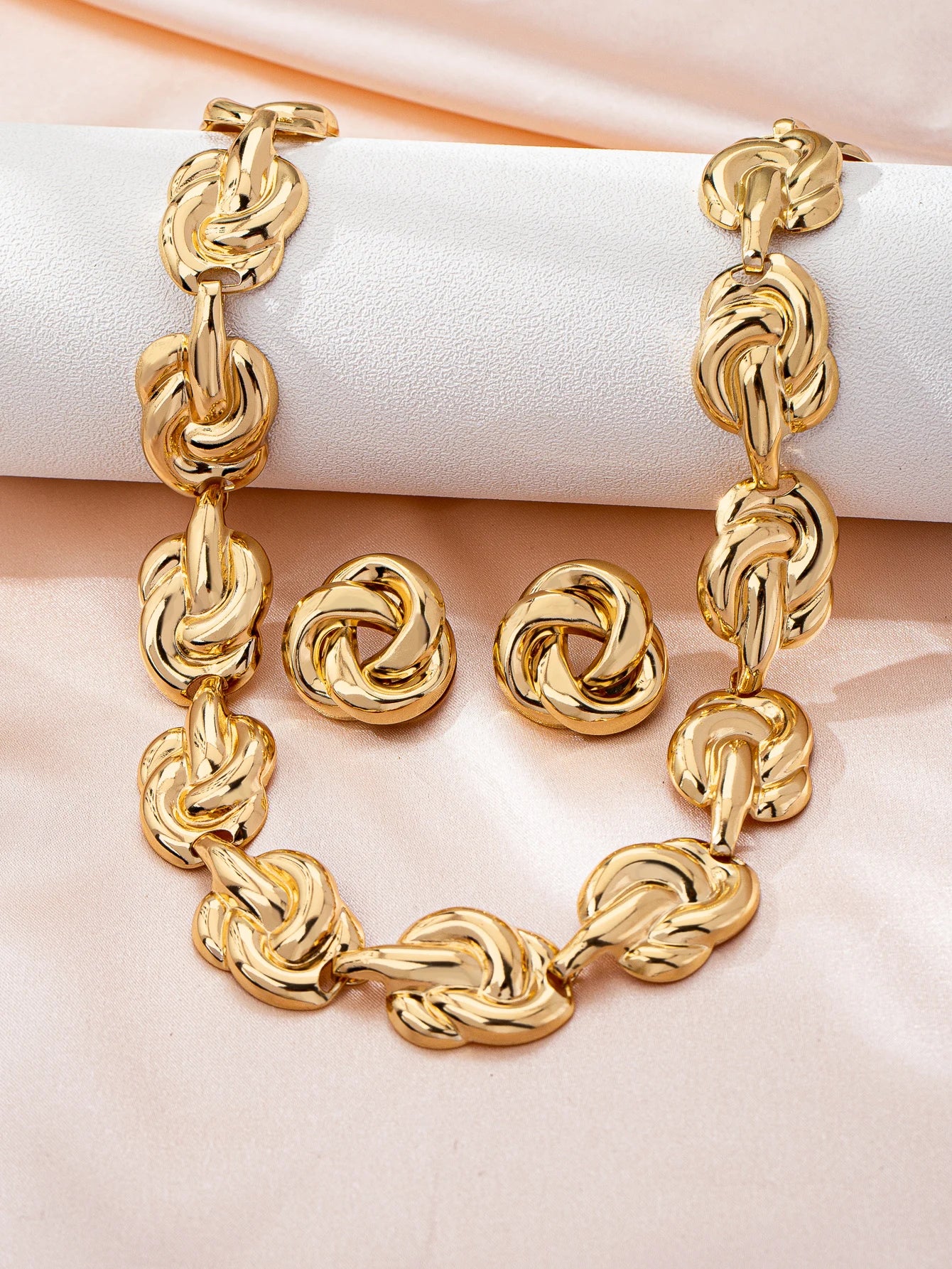 Gold chunky knot chain necklace and round stud earrings displayed on white roll on pink satin