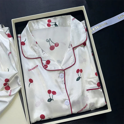 White pajama set with red cherry designs in a box on a dark background. Women’s cherries print summer pajamas set—faux satin V-neck top and shorts, funny cute style. Lightweight, comfortable, UK fashion set.