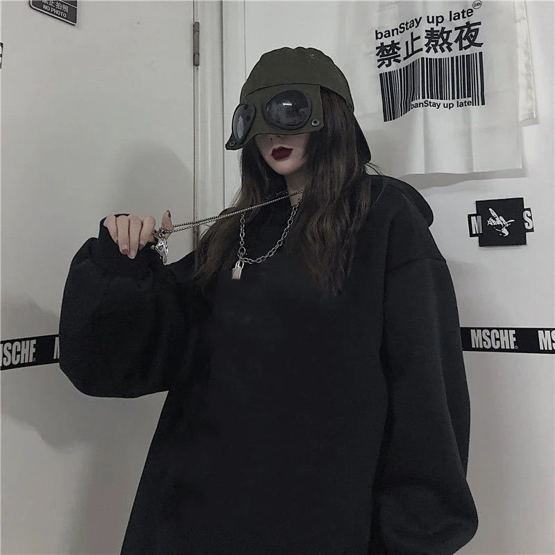 Person wearing a black hoodie and cap with goggles, standing against a white wall with text and logos.