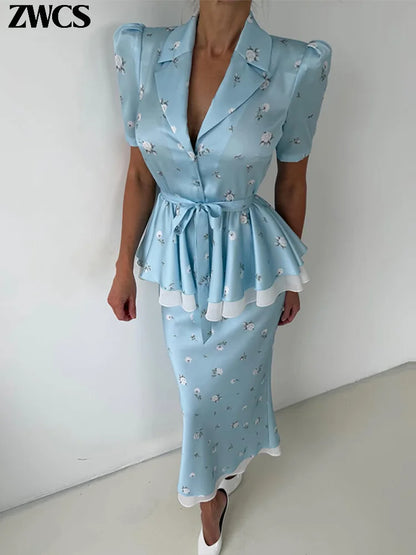 A woman modeling a light blue midi dress with a small white floral print, featuring a wrap-style collar, short puff sleeves, a tie waist, and a peplum hem with white ruffle trim. The brand name 'ZWCS' is visible in the top left corner.