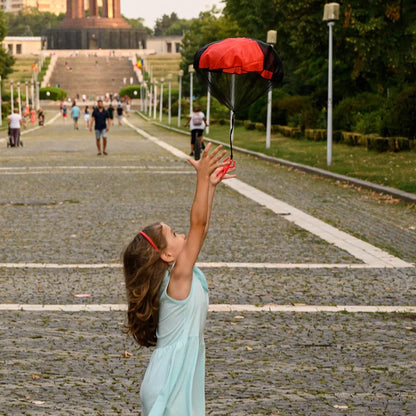 Child holding a red and black balloon in an outdoor setting with people and greenery. Colorful outdoor parachute toy for kids, tangle-free design. Durable plastic, unisex gift idea for boys and girls. UK delivery ftf fashion.