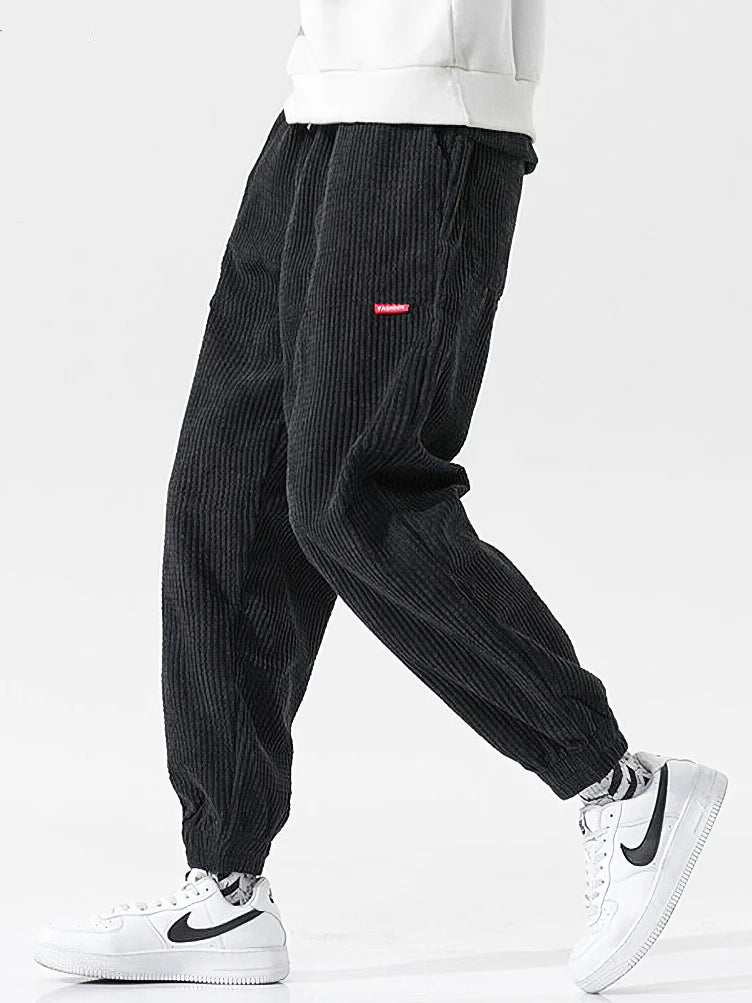 Black corduroy pants with a red label worn by a person on a white background. Men's baggy corduroy jogger pants, tech fleece sweatpants, autumn winter harem trousers, elastic waist, thermal, UK FTF Market.