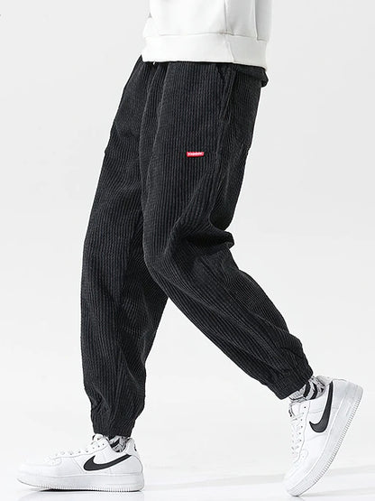 Black corduroy pants with a red label worn by a person on a white background. Men's baggy corduroy jogger pants, tech fleece sweatpants, autumn winter harem trousers, elastic waist, thermal, UK FTF Market.