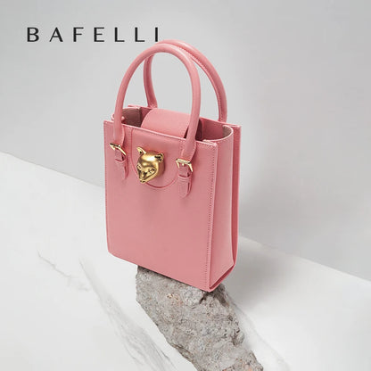 Pink handbag with gold accents on a light gray background, featuring the brand 'Bafelli'. Unisex mini tote crossbody bag in split leather with flap pocket and lock closure. Hard box shape and polyester lining. Casual versatile style.