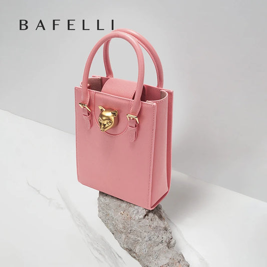 Pink handbag with gold accents on a light gray background, featuring the brand 'Bafelli'. Unisex mini tote crossbody bag in split leather with flap pocket and lock closure. Hard box shape and polyester lining. Casual versatile style.