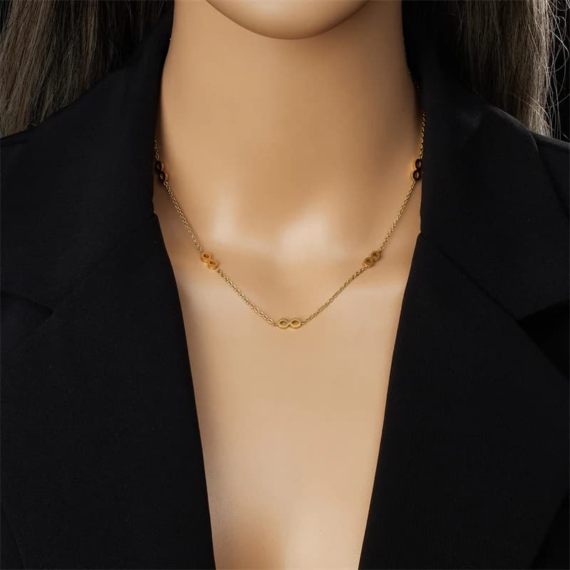 A gold infinity chain necklace with small flower-like charms, shown on a mannequin bust wearing a black blazer.