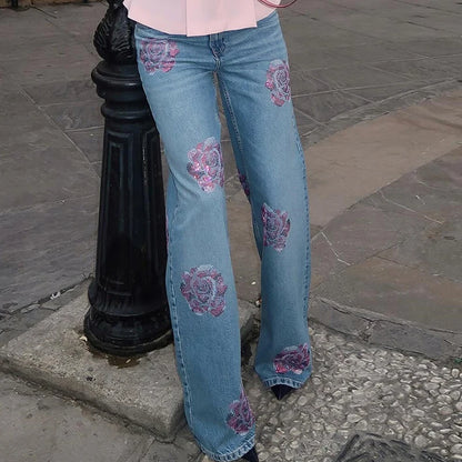 Person wearing blue jeans with floral embroidery on a stone pavement. Women’s denim jeans with flower rhinestones, loose wide leg, low waist, light wash. Stylish rhinestone casual trousers for UK fashion lovers.