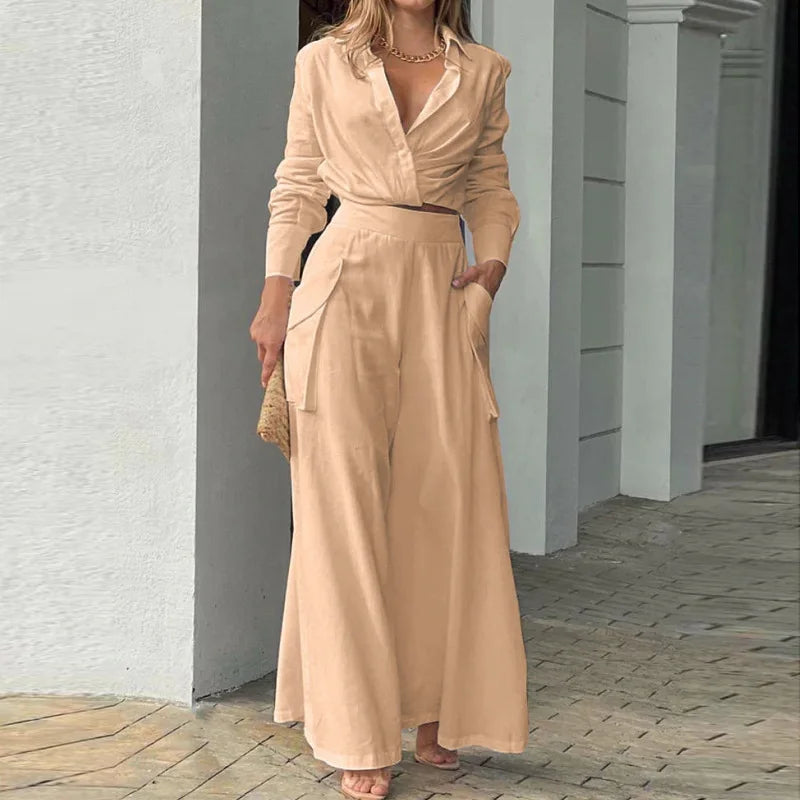 Woman wearing a beige outfit standing against a white wall. Women's elegant pants set with long sleeve crop shirt and high waist wide leg trousers. Office lady style. Solid, flattering, comfortable fit.