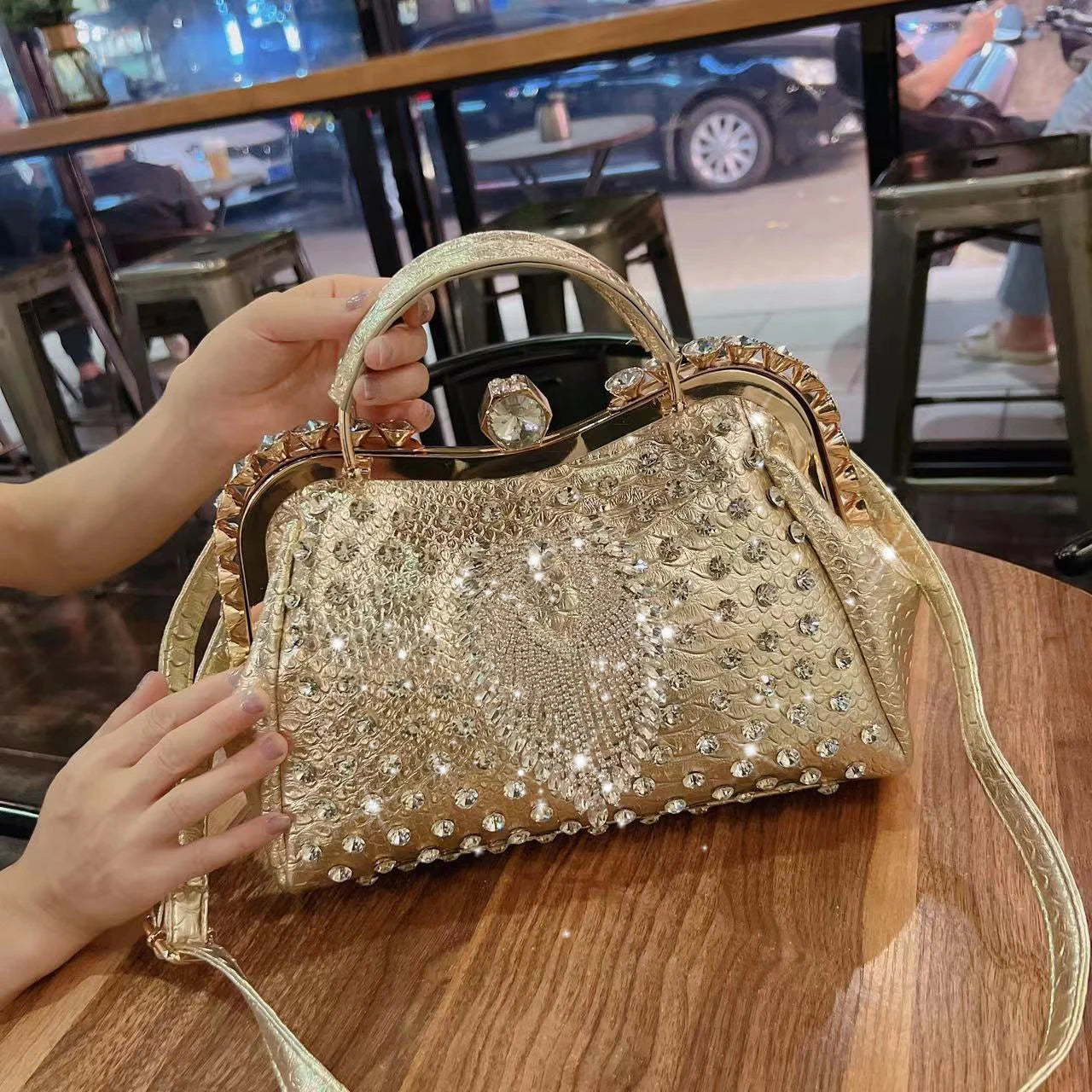 Gold embellished handbag on a wooden table with a blurred indoor background. Large genuine leather dumpling bag with diamond rhinestone clip and lock clasp. Soft handle, multiple compartments. Fashionable shoulder tote for women.