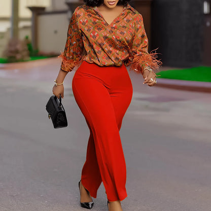 Woman in a patterned orange blouse and red pants walking outdoors. African print two-piece polyester outfit with satin printed shirt and high waist wide leg pants. Comfortable, elegant office wear for women.
