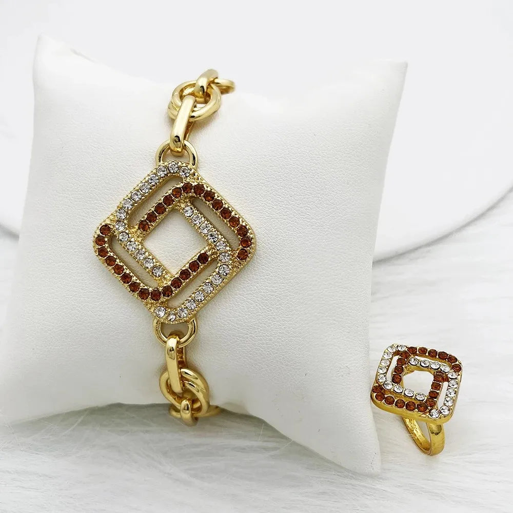 Gold-tone jewelry set featuring a bracelet and matching ring with a square geometric charm paved with clear rhinestones and garnished with small red stones, displayed on a white cushion.