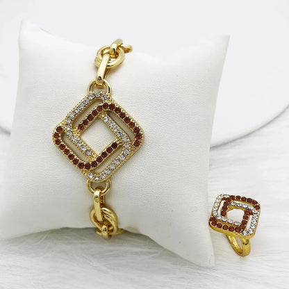 Gold-tone jewelry set featuring a bracelet and matching ring with a square geometric charm paved with clear rhinestones and garnished with small red stones, displayed on a white cushion.