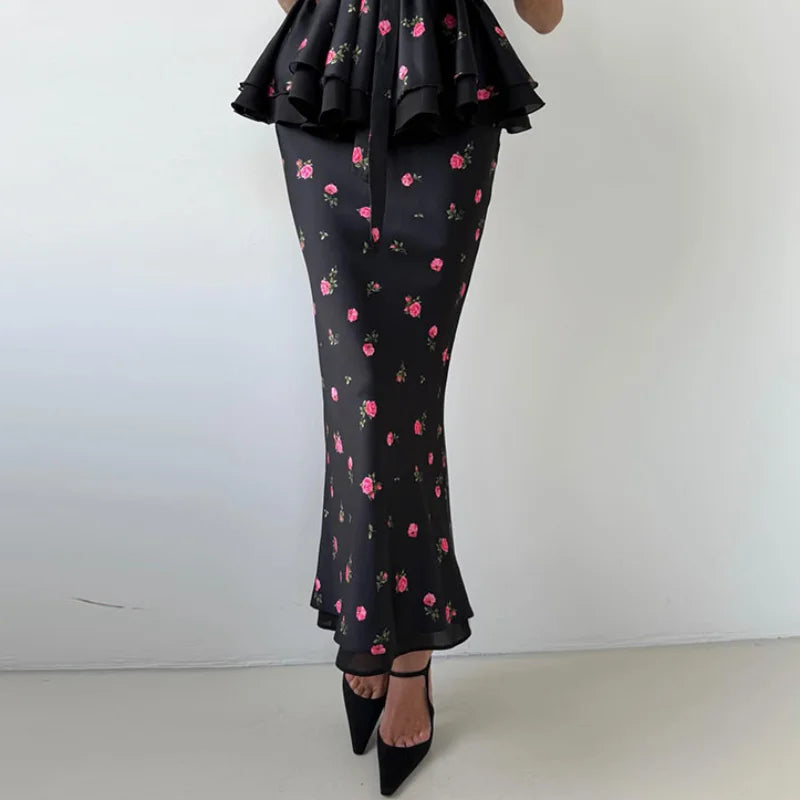 A model wearing a black ruffled maxi dress with a pink and green cherry blossom floral print, paired with black pointed-toe heels, shown from the waist down against a plain white wall.