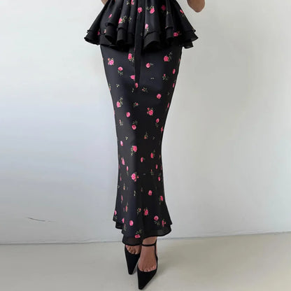 A model wearing a black ruffled maxi dress with a pink and green cherry blossom floral print, paired with black pointed-toe heels, shown from the waist down against a plain white wall.