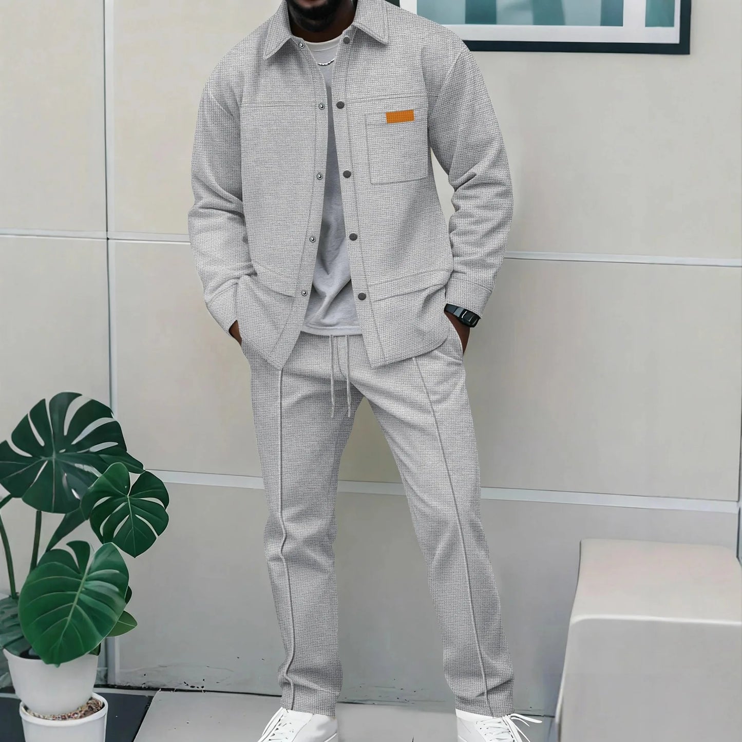 Person wearing a gray outfit with an orange patch, standing indoors next to a plant.
