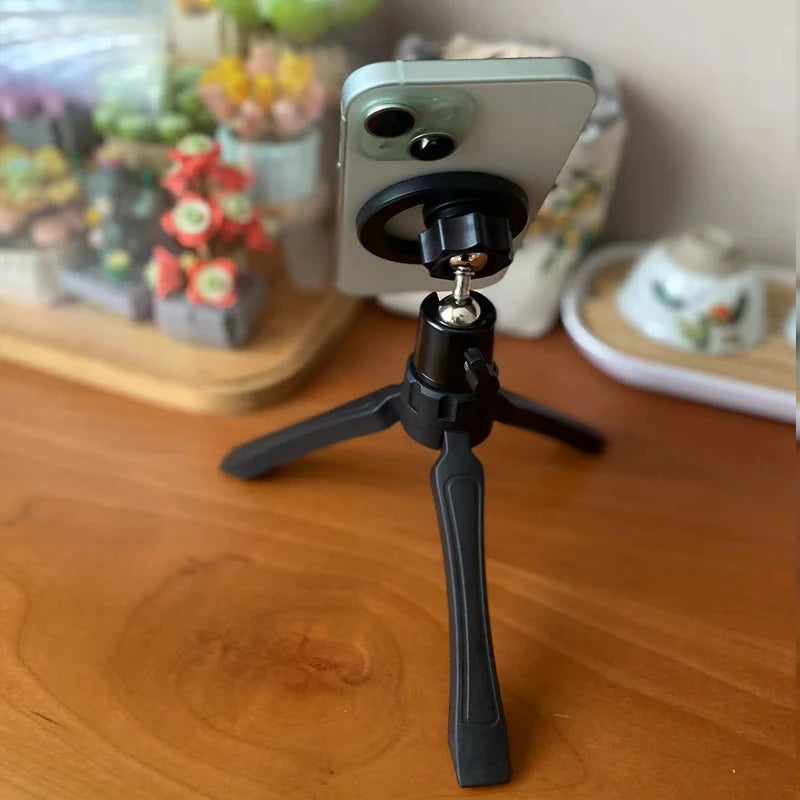Phone mounted on a small black tripod on a wooden surface with a blurred background. Magnetic phone tripod and wireless grip with remote control. Fits iPhone MagSafe and Samsung devices. Lightweight selfie stick tripod for creators.