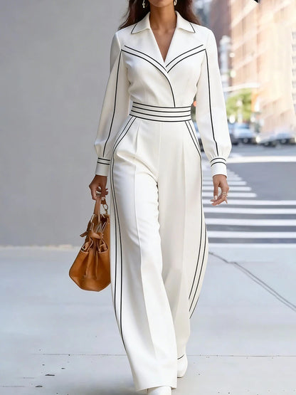 A woman walking on a city sidewalk wearing a white long-sleeve collared jumpsuit with black stripe trim details, holding a brown leather handbag.
