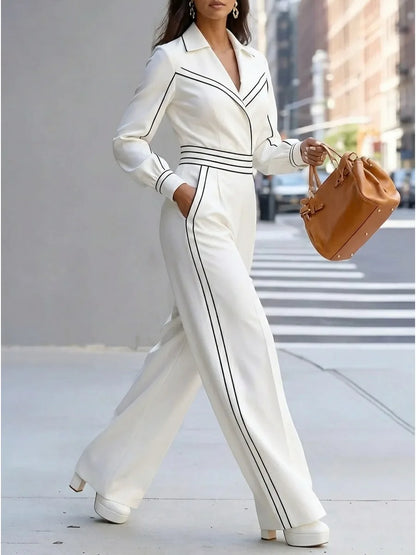 A woman walking on a city sidewalk wearing a white long-sleeve jumpsuit with black piping details, paired with white platform heels and holding a brown handbag.