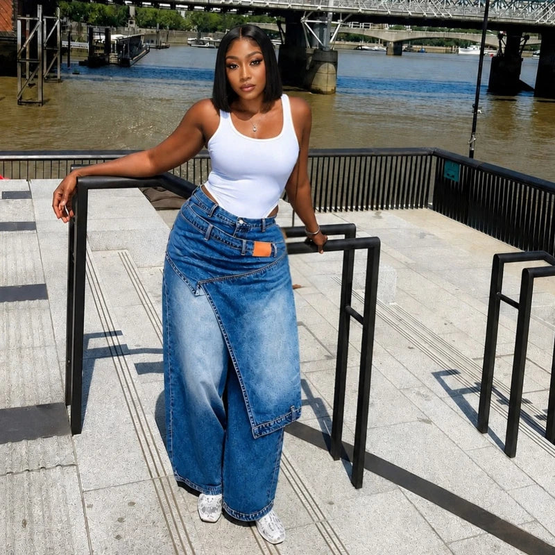 A woman posing outdoors on a riverside walkway, wearing a white ribbed tank top and a long, wide-leg blue denim skirt with a raw hem.