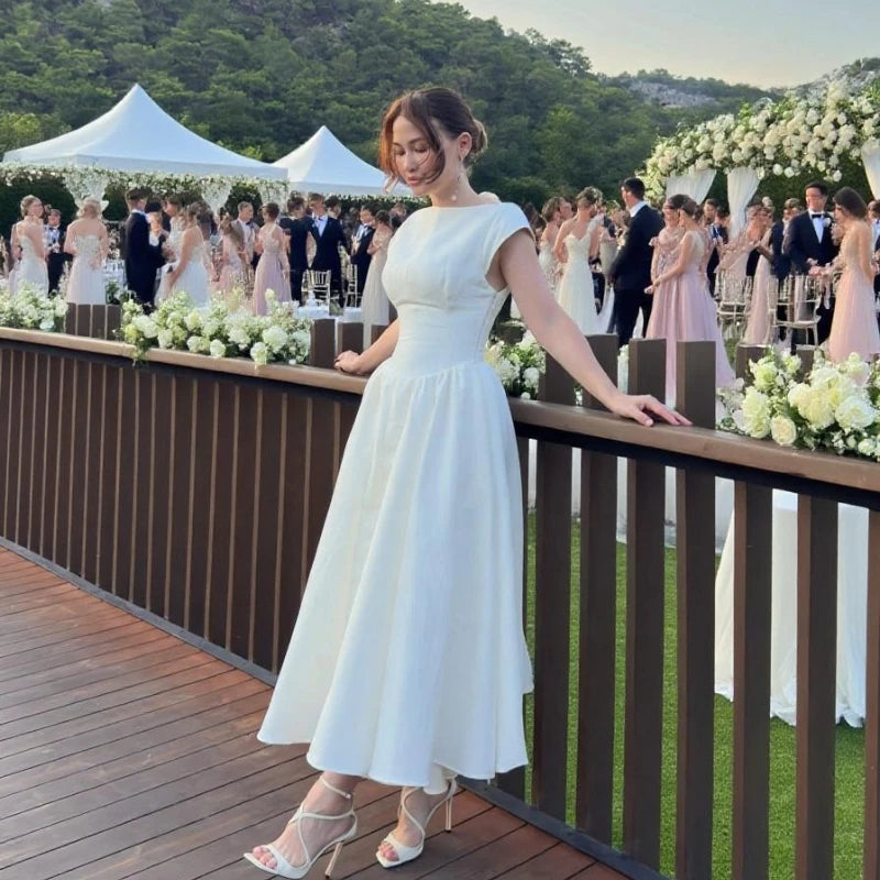 Woman in a white dress standing on a wooden deck with a wedding reception in the background.