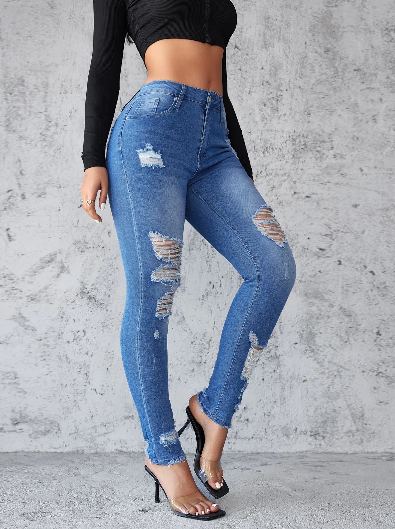 Person wearing blue ripped jeans against a gray concrete wall. Women’s high waist ankle length skinny jeans, ripped and distressed, pockets, solid slim pencil fit. Vintage streetwear by ftf fashion UK.