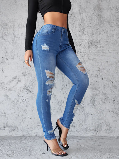 Person wearing blue ripped jeans against a gray concrete wall. Women’s high waist ankle length skinny jeans, ripped and distressed, pockets, solid slim pencil fit. Vintage streetwear by ftf fashion UK.
