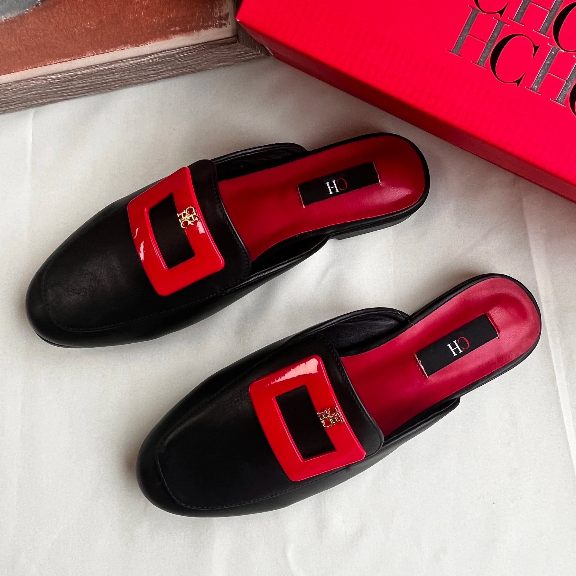 Black loafers with red buckles on a white surface, next to a red box.