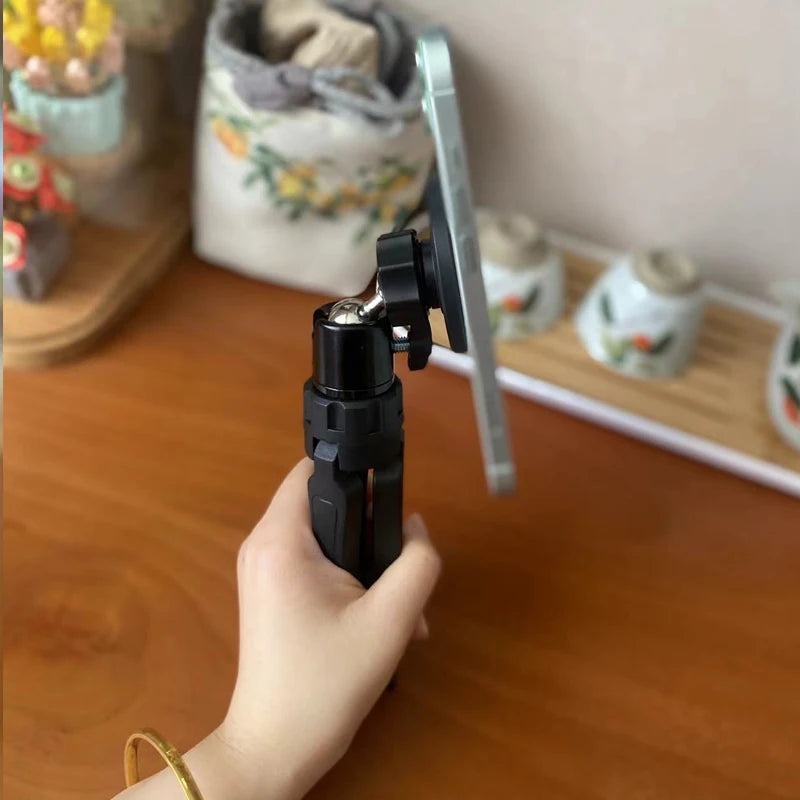 Hand holding a black suction cup mount on a wooden surface with decorative items in the background. Magnetic phone tripod and wireless grip with remote control. Fits iPhone MagSafe and Samsung devices. Lightweight selfie stick tripod for creators.