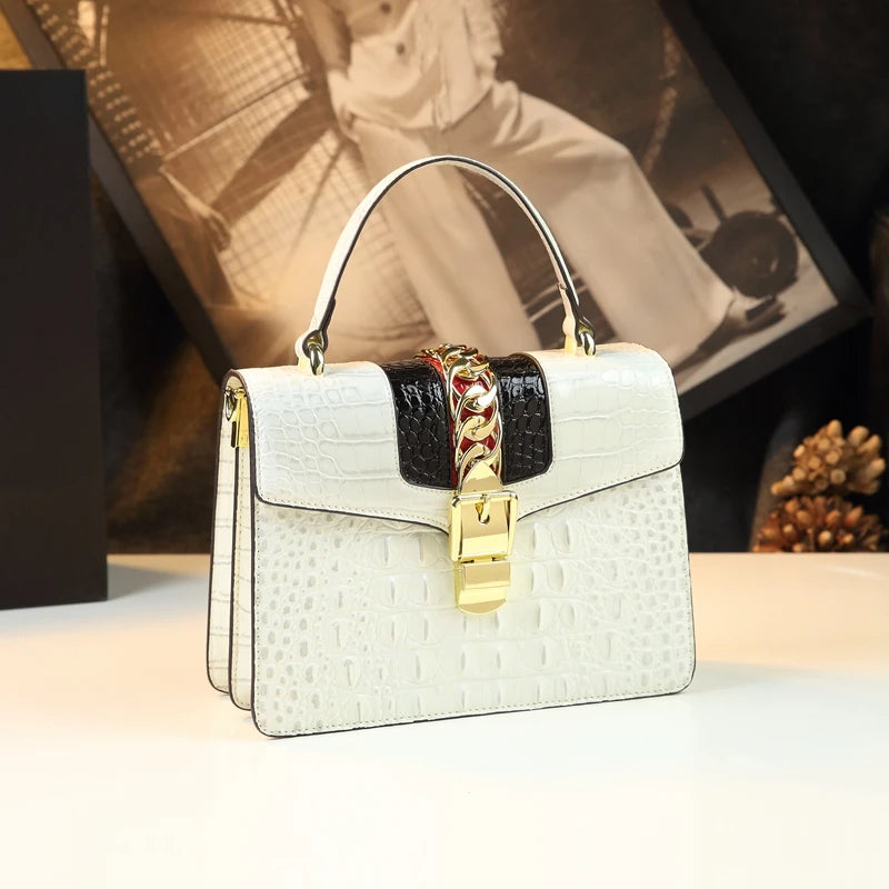 White handbag with gold chain and lock on a light surface with blurred background. Small genuine leather crocodile crossbody bag. Hard flap design with soft handle, zipper closure, and interior phone & compartment pockets.
