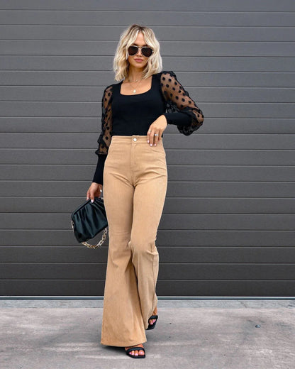 Woman wearing a black top with sheer sleeves and beige pants against a gray wall. Slim micro flare corduroy trousers with elastic waistband, screw thread button detail, casual style for women. Available in multiple colors UK FTF Market.