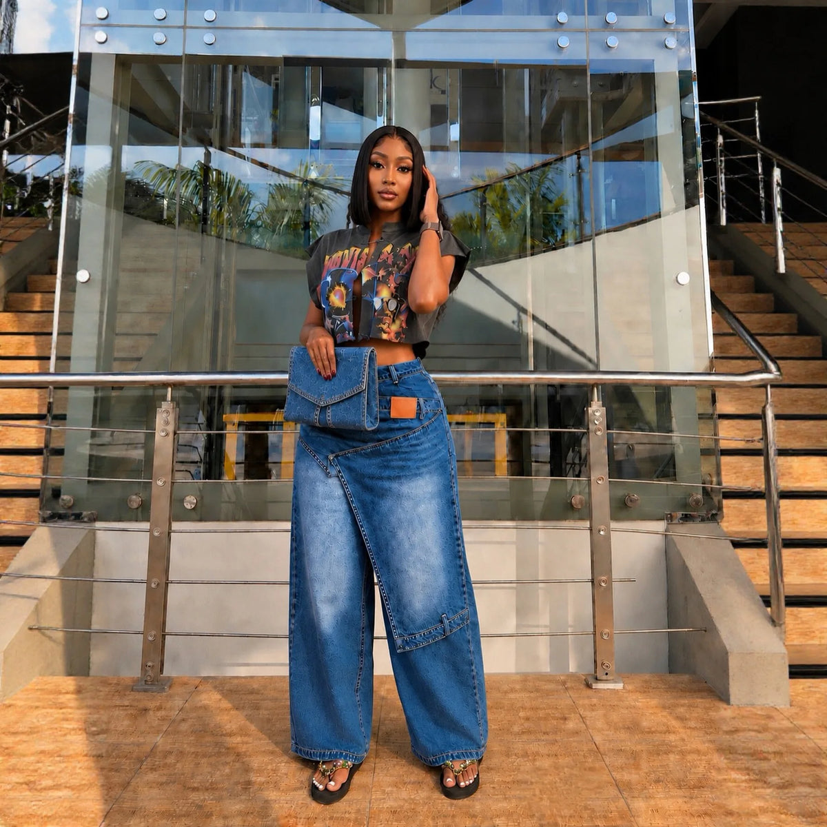 A woman stands in front of a modern glass building with stairs, wearing a black graphic crop top, a blue denim wrap-style skirt with a front flap pocket, and black sandals.