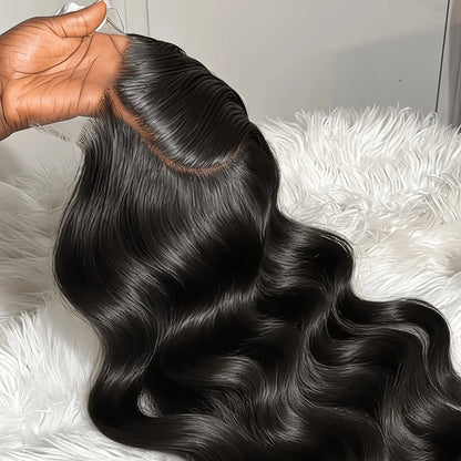 A close-up of a long, wavy black hair extension or wig being held by a hand, showing the hair texture and a natural-looking root line, set against a white faux fur background.