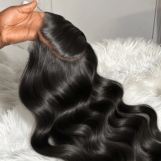 A close-up of a long, wavy black hair extension or wig being held by a hand, showing the hair texture and a natural-looking root line, set against a white faux fur background.