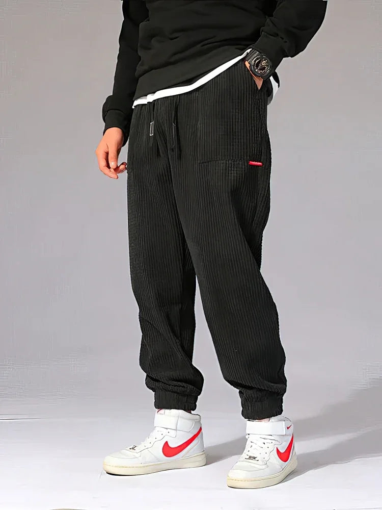 Person wearing black corduroy jogger pants with white sneakers featuring red accents on a gray background. Men's baggy corduroy jogger pants, tech fleece sweatpants, autumn winter harem trousers, elastic waist, thermal, UK FTF Market.