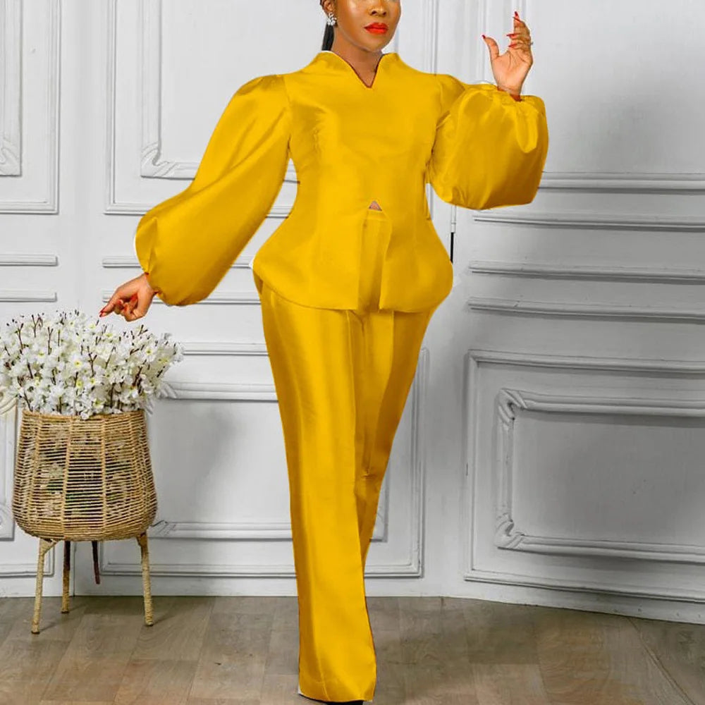 Woman in a yellow outfit standing next to a decorative basket with flowers against a white paneled wall. Women's 2-piece pants set with solid satin blouse, straight high-waist trousers. Office lady luxury outfit for professional and elegant occasions.