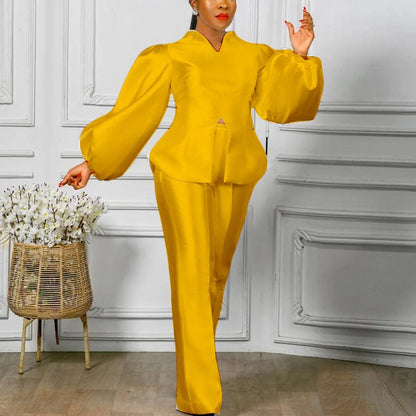 Woman in a yellow outfit standing next to a decorative basket with flowers against a white paneled wall. Women's 2-piece pants set with solid satin blouse, straight high-waist trousers. Office lady luxury outfit for professional and elegant occasions.