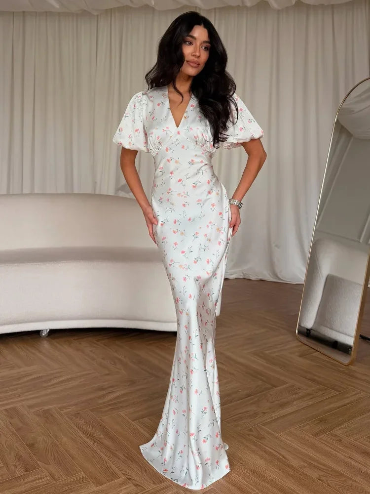 A woman models a white wrap-style maxi dress with a delicate pink and green floral print, featuring short flutter sleeves and a V-neckline, set against a neutral indoor backdrop with a cream sofa and wooden floor.
