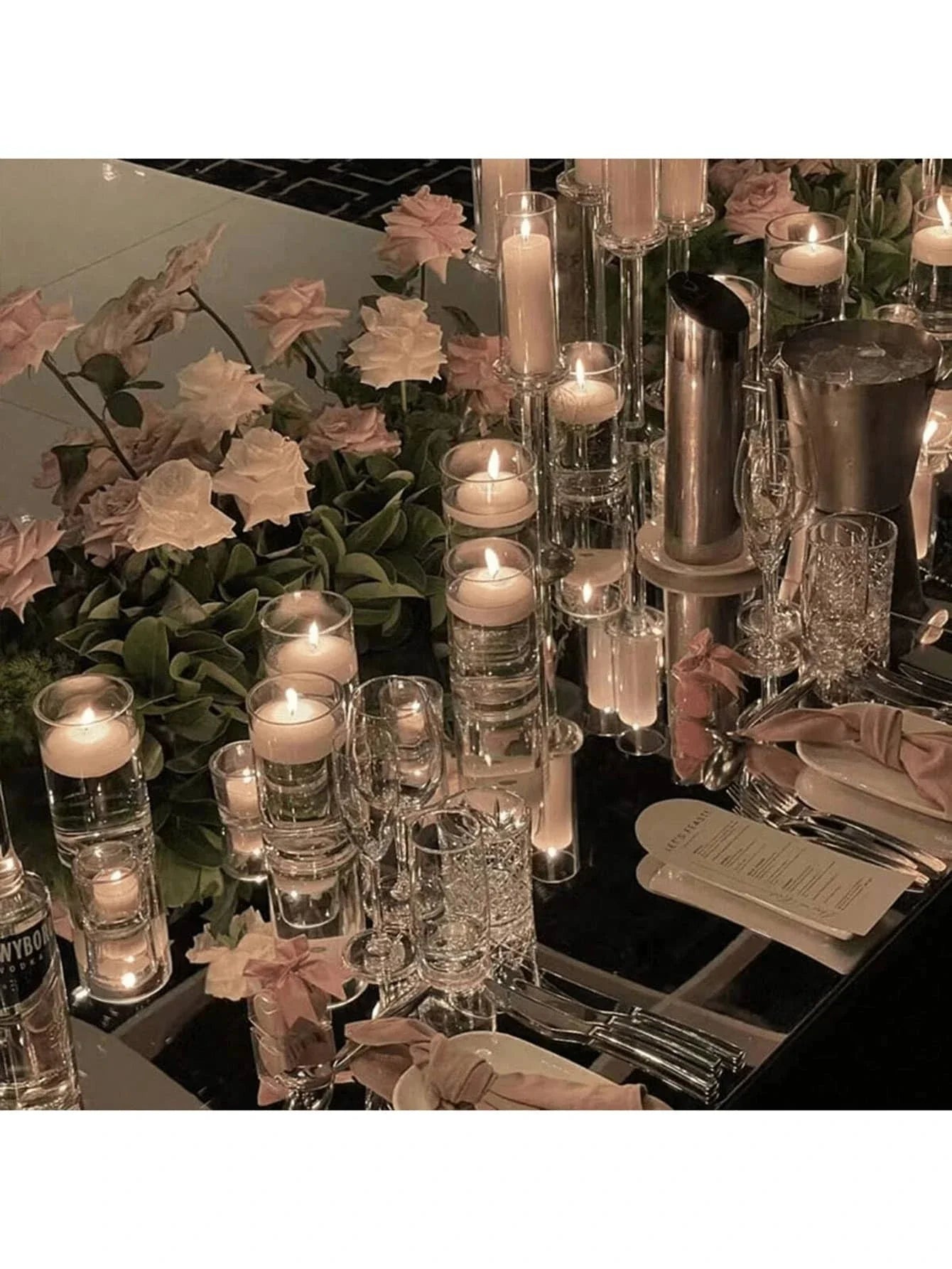 Elegant table setting with candles, glasses, and flowers on a reflective surface.