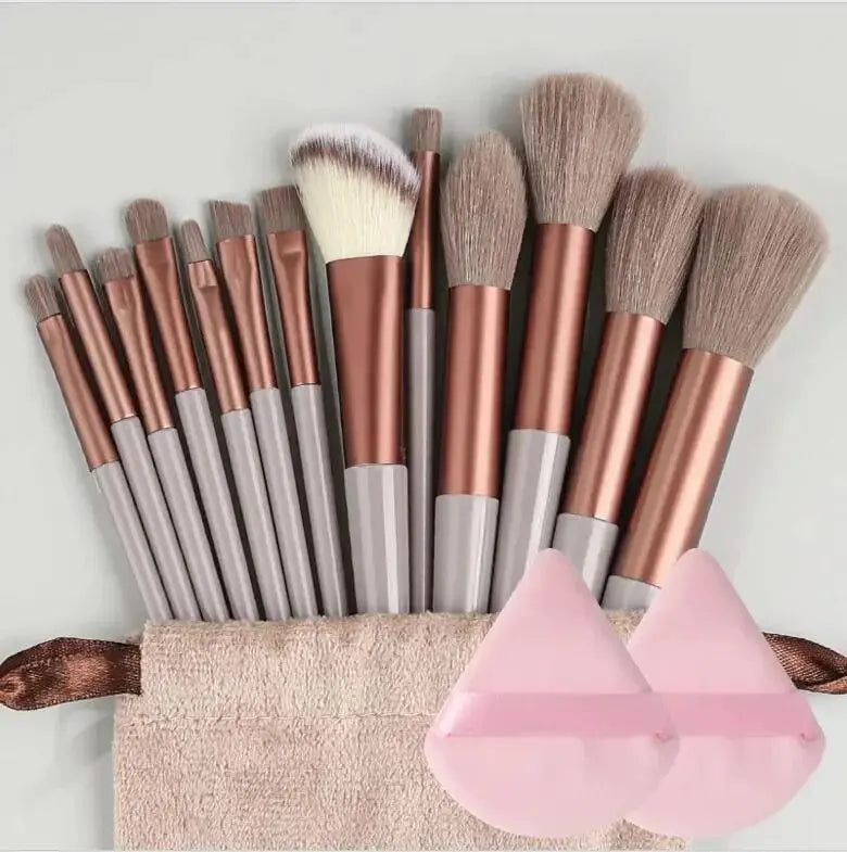 Set of makeup brushes with rose gold and silver handles, a beige pouch, and two pink silicone sponges on a light gray background. Soft fluffy makeup brush set with wool fiber bristles and plastic handles. Complete set for powder, foundation, eyeshadow, and beauty blending.