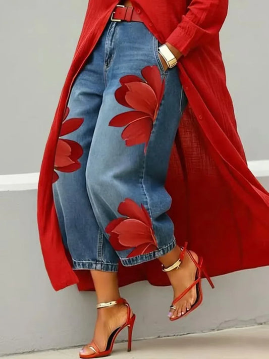 A person wearing blue cropped jeans with large red floral applique patches, a flowing red cardigan, and red high-heeled sandals, standing against a light gray wall.