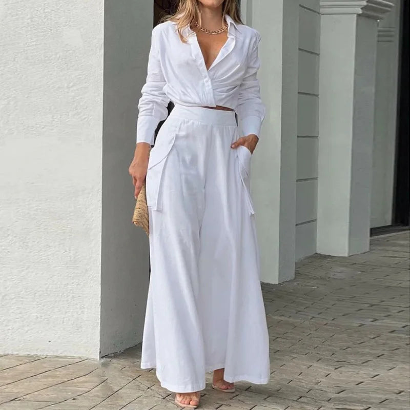 Woman wearing a white outfit standing against a light-colored wall. Women's elegant pants set with long sleeve crop shirt and high waist wide leg trousers. Office lady style. Solid, flattering, comfortable fit.