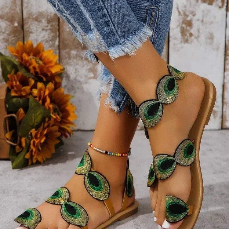 Beige sandals with peacock feather designs worn with denim shorts, against a rustic wooden background.