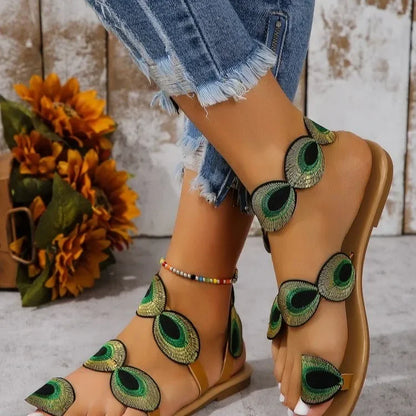 Beige sandals with peacock feather designs worn with denim shorts, against a rustic wooden background.