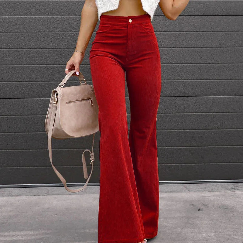 Person wearing red pants holding a beige handbag against a gray wall. Slim micro flare corduroy trousers with elastic waistband, screw thread button detail, casual style for women. Available in multiple colors UK FTF Market.